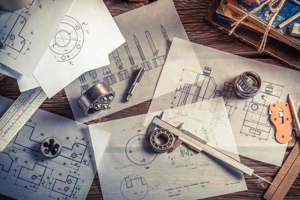 Five detailed engineering specification drawings laid on a table along with an engineering scale and bearings