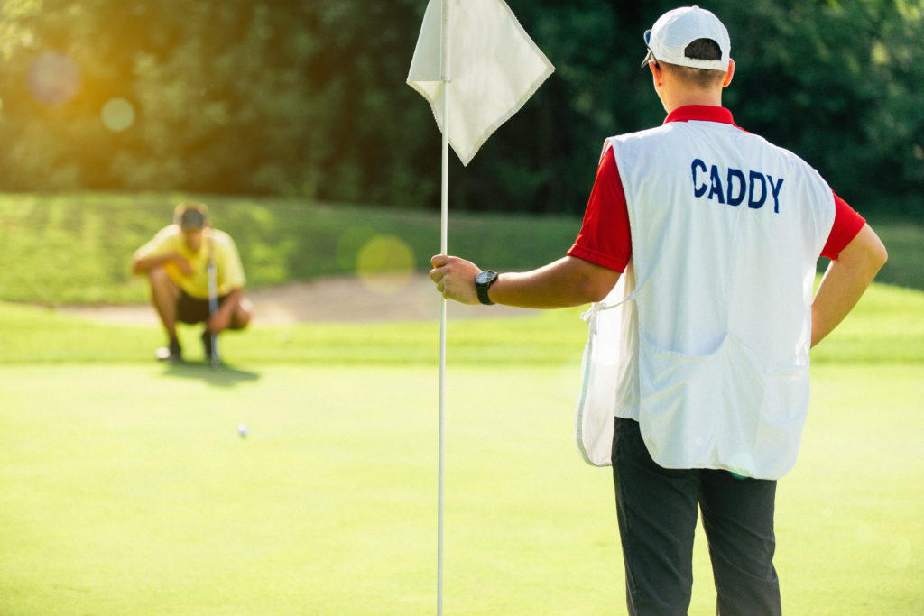 boy caddie, hoping to get an outside scholarship for caddying, holding flag over hole on golf course. Man in distance crouching to look to see if he can make the shot. 
